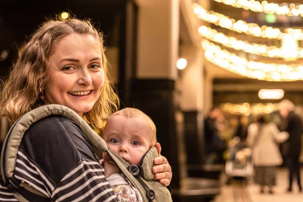 A woman stands in the Living Worlds Gallery at Manchester Museum, smiling while holding a baby in a sling strapped to her front.