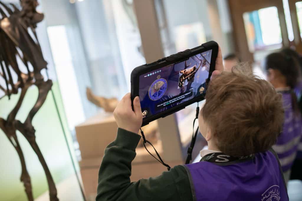 A primary school pupil wearing a purple tabard over their uniform uses a tablet computer to take a picture of an animal skeleton in a museum.