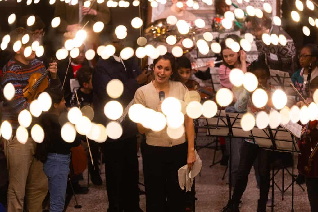 A view through fairy lights of one of the Olympias Music Foundation teachers speaking at their Christmas concert in Manchester Museum's Living Worlds gallery.